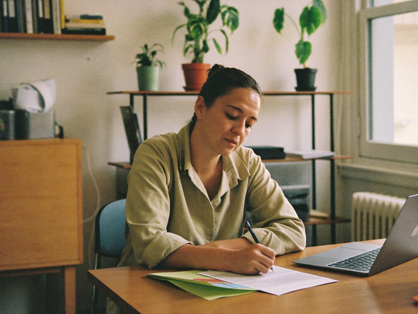 A woman writing at a desk in a light-filled community office, laptop and printed pages in front of her.
